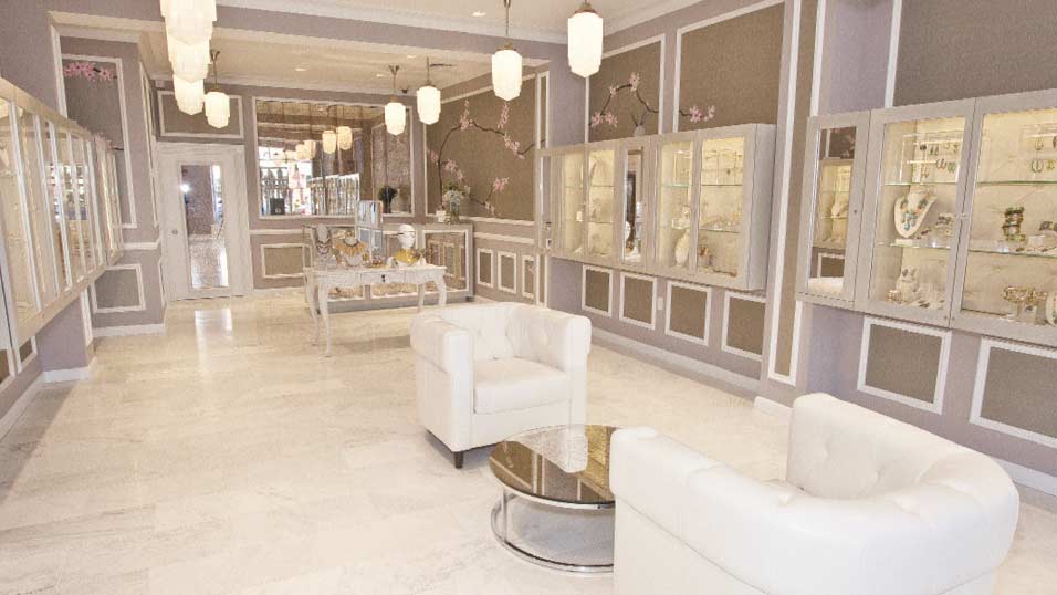 A wide interior view of the luxury Alexis Bittar retail showroom built by MOMA Construction Group, featuring light gray paneled walls with elegant crown molding, built-in glass display cases showcasing jewelry, plush white tufted armchairs, a marble-topped display table, and contemporary pendant lighting.