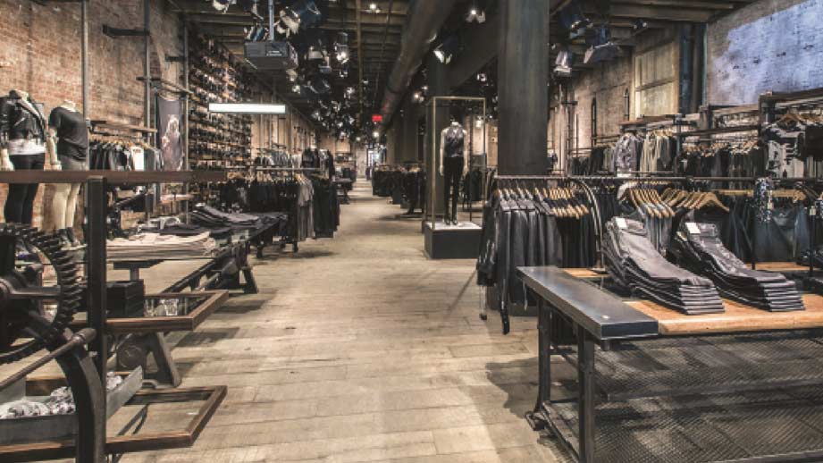 A wide interior view of the AllSaints retail showroom built by MOMA Construction Group, showcasing an industrial design with exposed brick walls, polished concrete flooring, a wooden ceiling with track lighting, and custom dark metal clothing racks displaying apparel.
