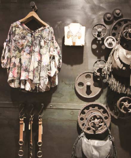 A detailed view of a rustic and industrial retail display wall built by MOMA Construction Group for the AllSaints showroom, featuring a floral print blouse on a wooden hanger, a designer purse, and a unique background decorated with various sizes of antique metallic industrial gears and wheels.