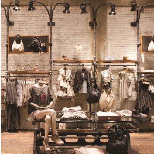 An interior view of the AllSaints retail showroom built by MOMA Construction Group, featuring an industrial aesthetic with exposed piping and brick, a mannequin showcasing a patterned dress and boots, and custom hanging clothing racks with layered apparel.
