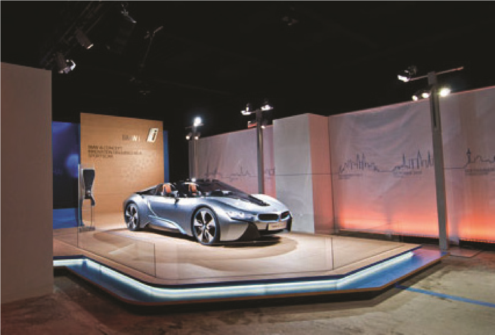 A high-end commercial vehicle showroom built by MOMA Construction Group for the BMWi line, featuring an electric sports coupe displayed on an elevated, illuminated wooden platform surrounded by architectural glass railings and vibrant accent wall lighting.