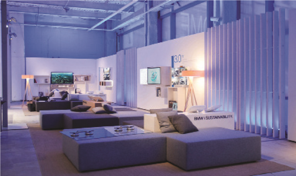 A modern, minimalist interior view of the BMWi showroom lounge built by MOMA Construction Group, featuring sleek modular gray seating, light wood flooring, contemporary floor lamps, and futuristic blue and white ambient accent lighting.