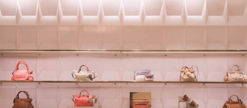 A wide interior view of a luxury retail display built by MOMA Construction Group for the Chloé boutique, showcasing multiple designer leather handbags elegantly arranged on clean, minimalist blush pink shelving with gold metal trim and warm backlight illumination.