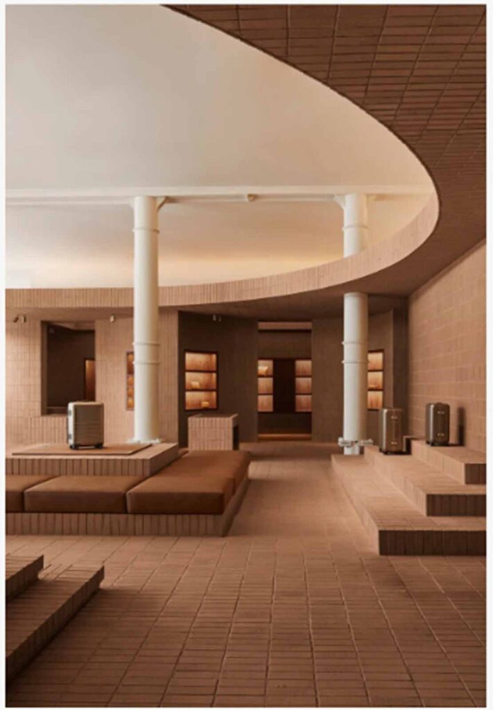 A wide, low-angle interior view of a modern multi-level retail showroom in Soho featuring a large curved mezzanine balcony, custom terracotta brick tile flooring, and integrated plush bench seating built into stepped platforms.