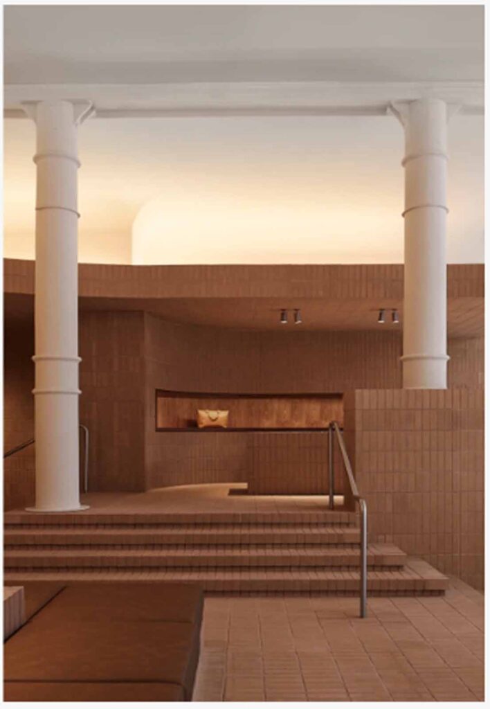 A multi-level retail showroom interior in Soho featuring terracotta-colored brick tile steps leading to an elevated floor section with two prominent white structural pillars.