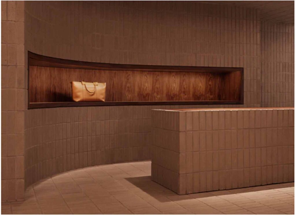 A modern retail showroom interior in Soho featuring a curved terracotta brick-tiled wall with an illuminated, recessed wooden display shelf holding a luxury tan leather handbag.