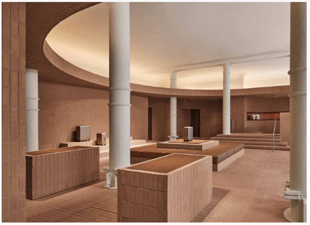 A wide interior view of a modern multi-level retail showroom in Soho featuring terracotta-colored brick tile flooring, custom display blocks, prominent white structural columns, and an integrated amphitheater-style seating layout.
