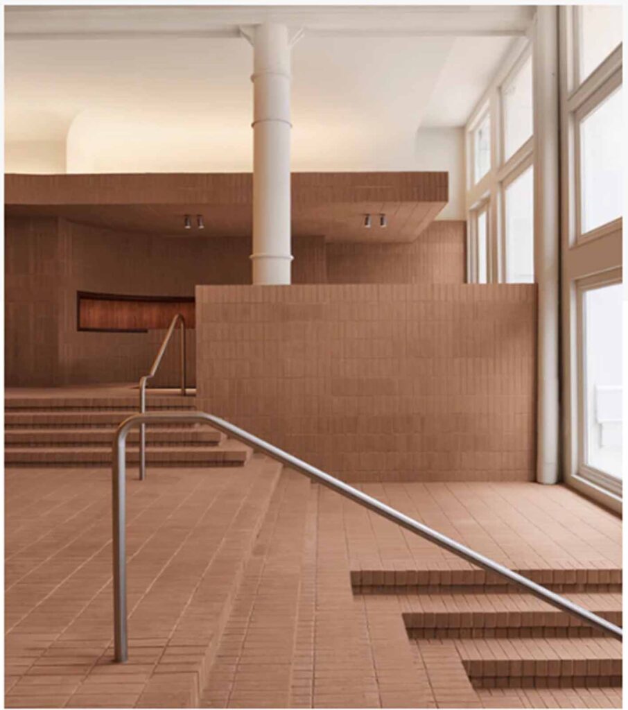 A modern multi-level retail showroom interior in Soho featuring terracotta-colored brick tile stairs, custom matching brick-wrapped walls, and a sleek stainless steel handrail leading to an upper mezzanine level.
