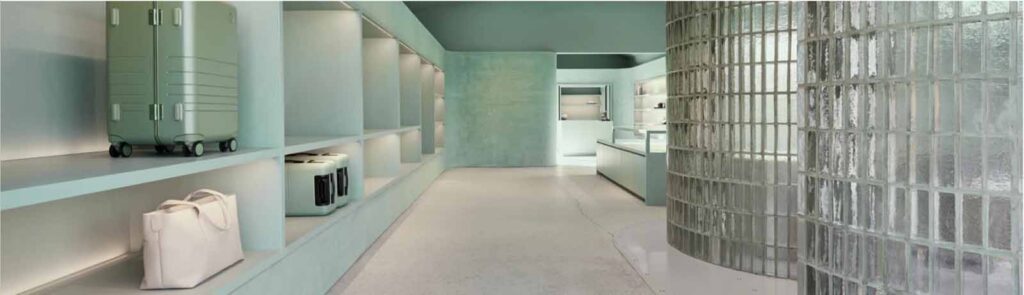 A wide interior view of the Monos showroom on Abbot Kinney featuring a long curved glass-block wall, light teal built-in shelving displaying luggage, and a concrete floor.