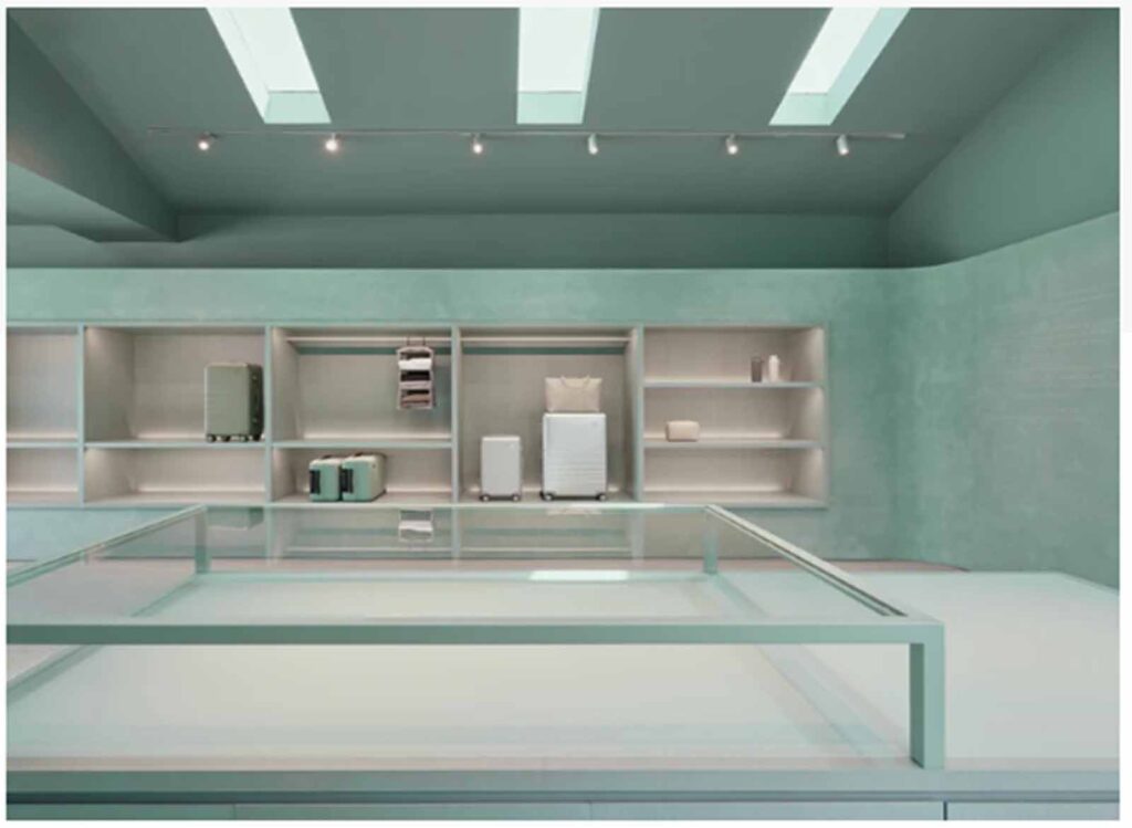 A modern boutique interior on Abbot Kinney featuring a sleek glass display case in the foreground and minimalist built-in wall shelving displaying luxury luggage and travel cases.