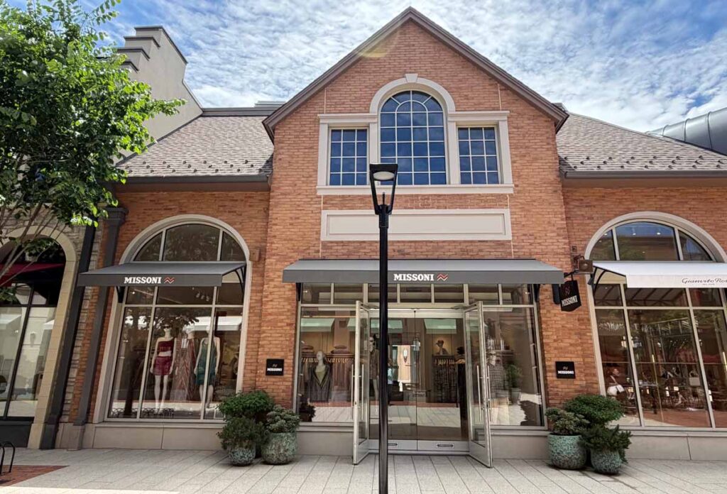 The classic brick commercial storefront entrance of Missoni luxury retail boutique at Belmont Park, managed by MOMA Construction Group, featuring prominent arched windows, sleek dark fabric awnings over structural glass entry doors, and manicured decorative planters.