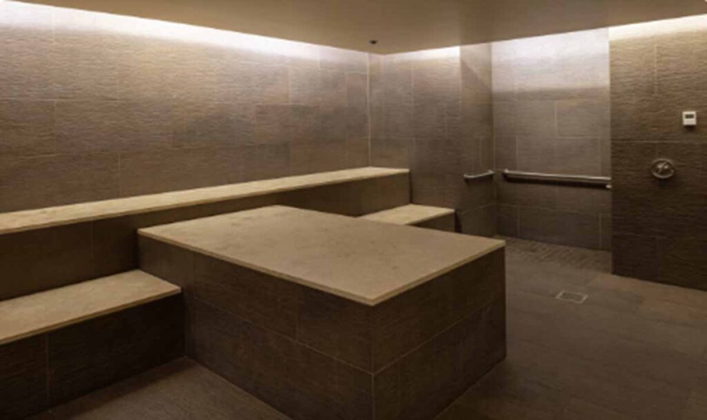 A luxury commercial steam room or thermal suite inside an Exhale wellness medspa, featuring dark tile walls and large stone slab benches under soft recessed ambient lighting.