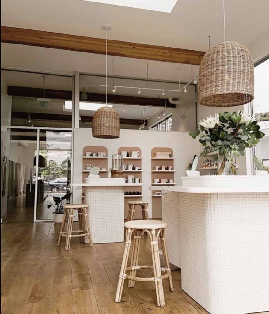 A bright showroom and reception area iniside a Heyday wellness medspa in Silver Lake, featuring white-tiled checkout counters with light wood tops, matching woven cane stools, industrial wood ceiling beams, and large basket pendant light fixtures.