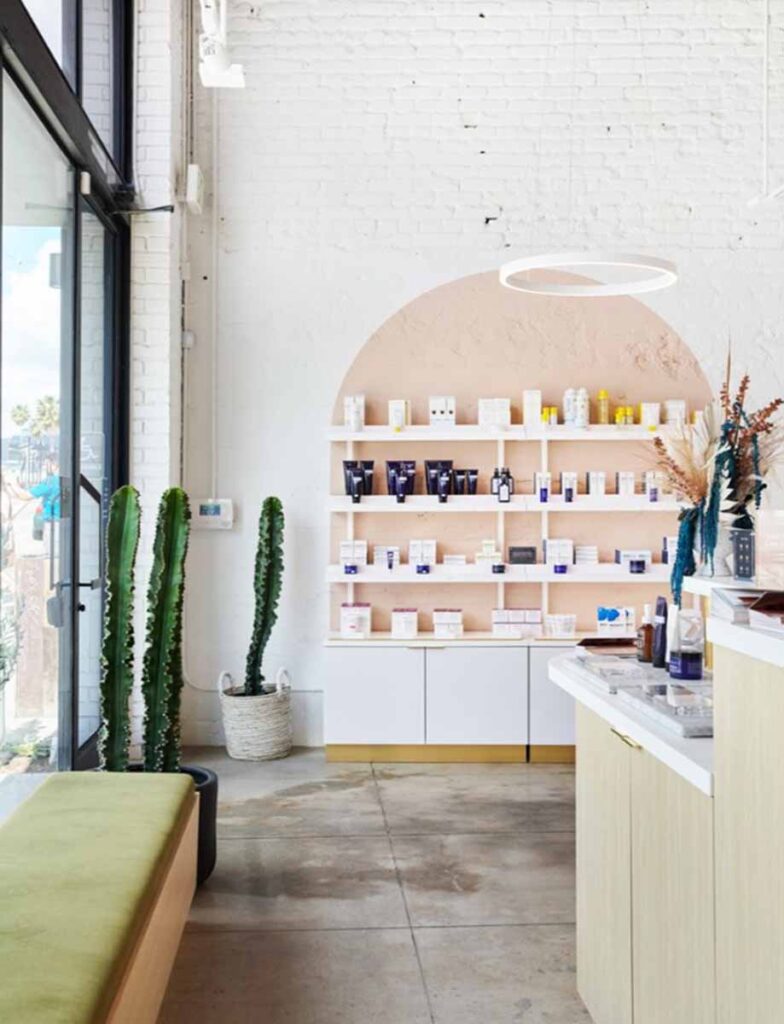 A contemporary retail display area inside a Heyday wellness medspa in Silver Lake. Featuring a large arched alcove wall painted in a soft terracotta hue, built-in white product shelves stocked with skincare items, and a white minimalist counter with gold hardware.