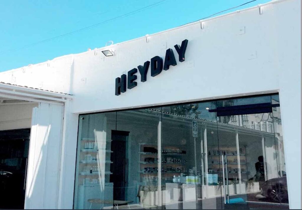 The exterior storefront facade of a Heyday wellness medspa in NoHo featuring a clean white building exterior, a large bold black Heyday sign, and expansive structural glass windows revealing the modern retail showroom interior.