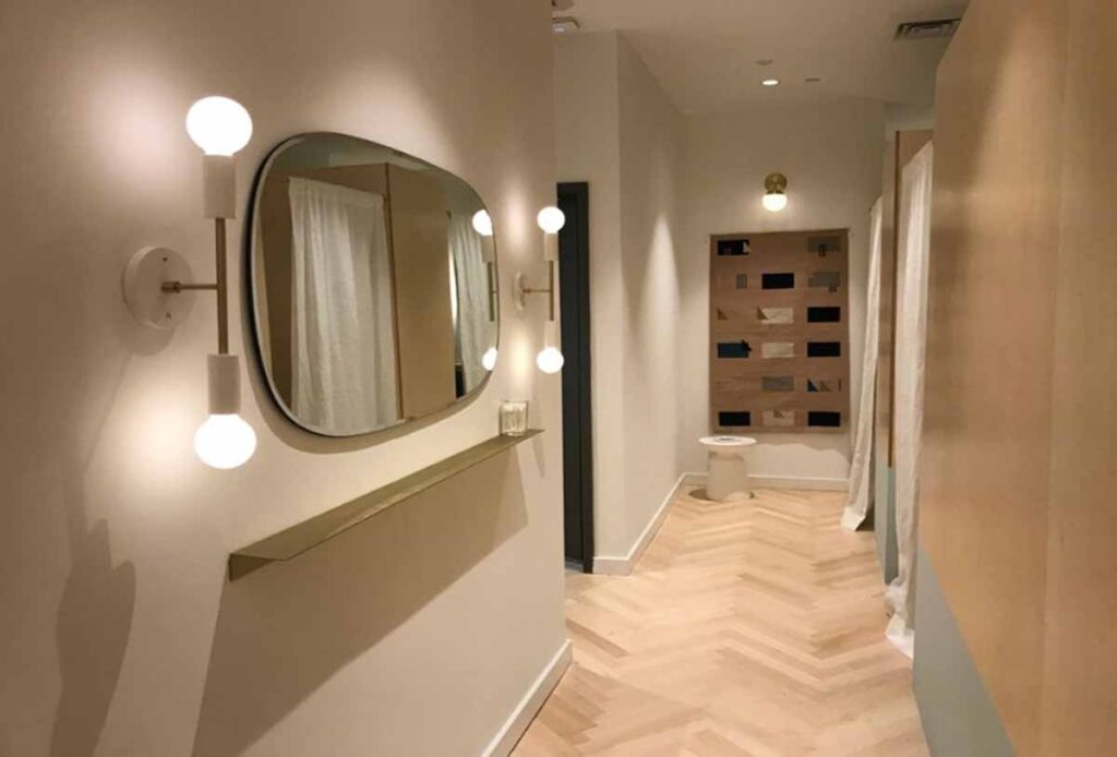 A modern vanity station inside a NoHo wellness medspa featuring an asymmetrical oblong mirror with a brushed gold shelf, flanked by two vertical minimalist globe sconces on a light herringbone wood floor corridor.