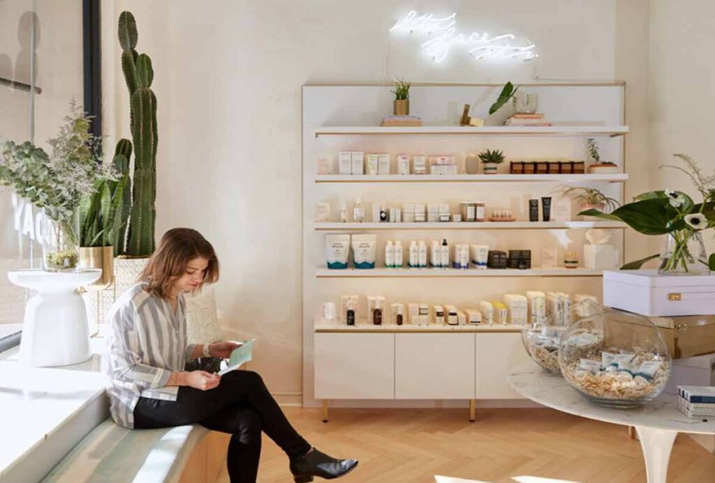 A woman sits on a built-in wooden bench reading a menu inside a bright Heyday wellness medspa in NoHo. In the background, a large white retail display unity is stocked wiht skincare products beneath an illuminated neon sign, alongside a marble display table.