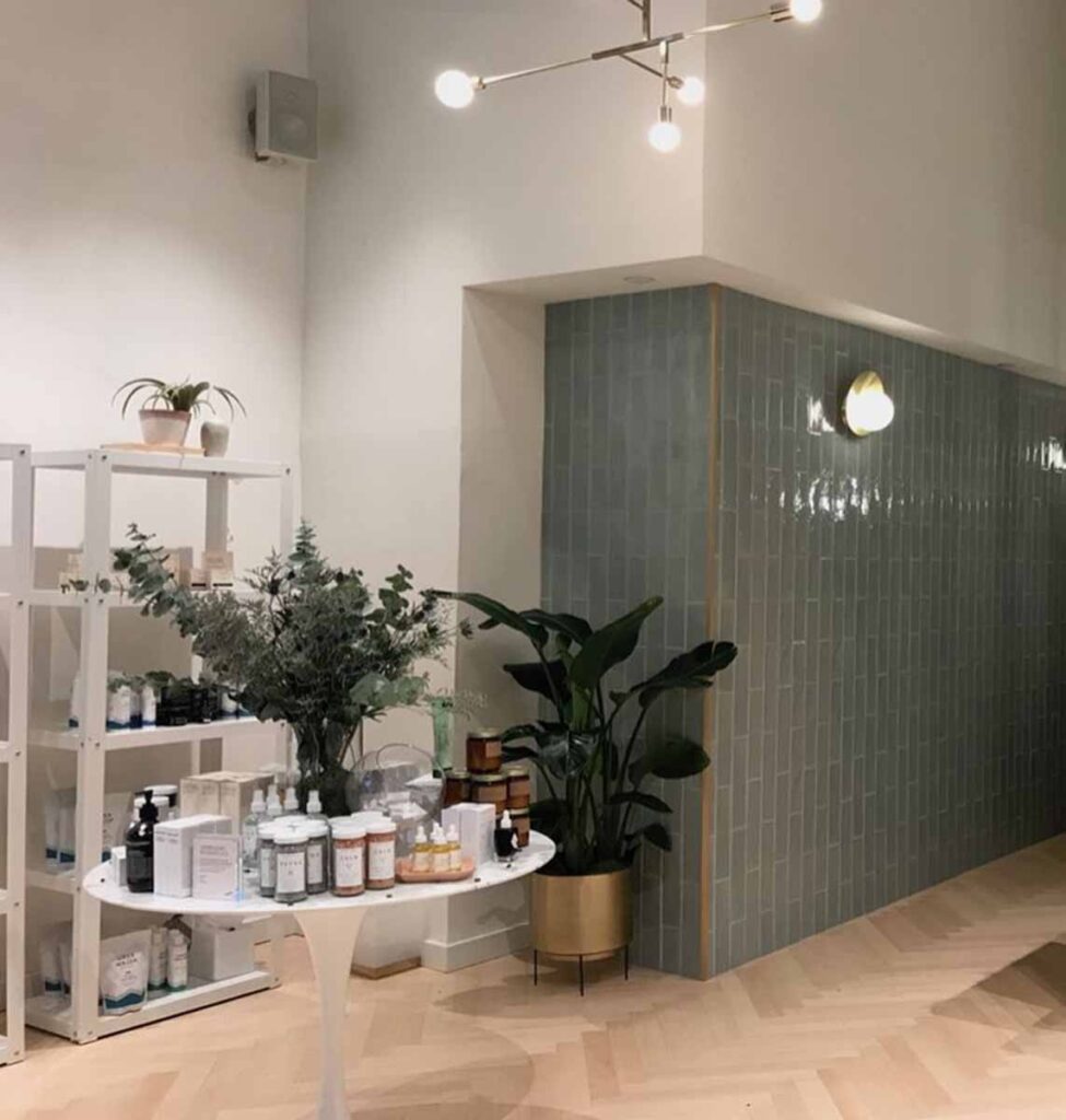 A contemporary wellness medspa or skincare retail showroom featuring a curved white product display table and an accent wall finished in vertical grey-green subway tiles under modern track lighting.