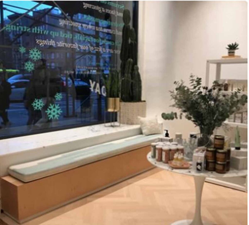 A modern, welcoming lobby and waiting area inside a NoHo wellness medspa featuring a custom built-in wooden bench with a light cushion, a round white display table showcasing skincare products and a vase of eucalyptus, and large street-facing windows with seasonal snowflake decals.