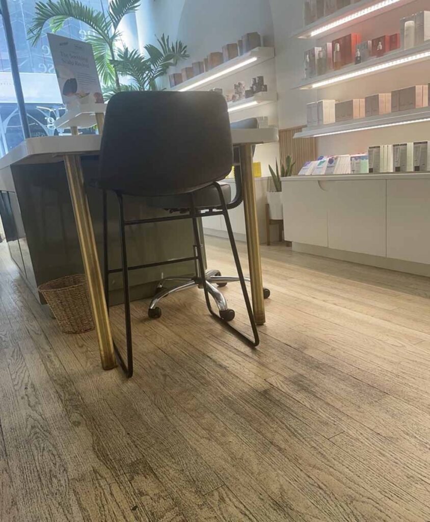 Commercial medical spa showroom interior with integrated under-cabinet LED strip lighting, a hardwood plank floor, and a modern black leather counter stool at a white laminate product display island.