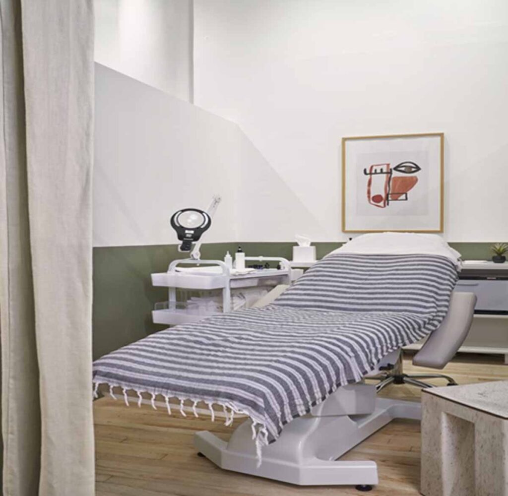 Commercial medical spa treatment room featuring an adjustable white anesthetic facial bed with a striped blanket, textured quarz terrazzo side tables and two-tone minimalist wall with fine art framing.