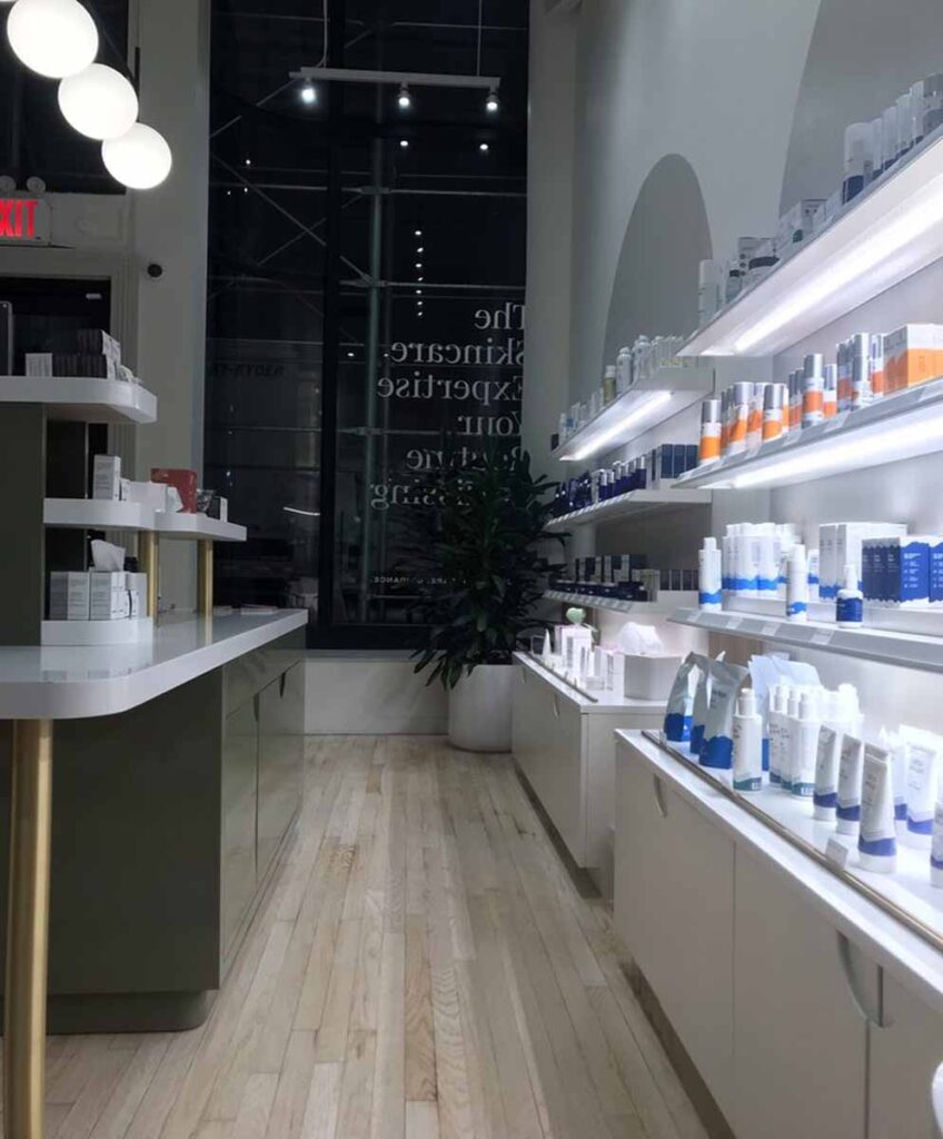 Commercial medical spa beauty boutique retail showroom renovation with dual-level product shelving and integrated under-shelf linear LED lighting, light hardwood floors, and a view through a lare glass display storefront window.