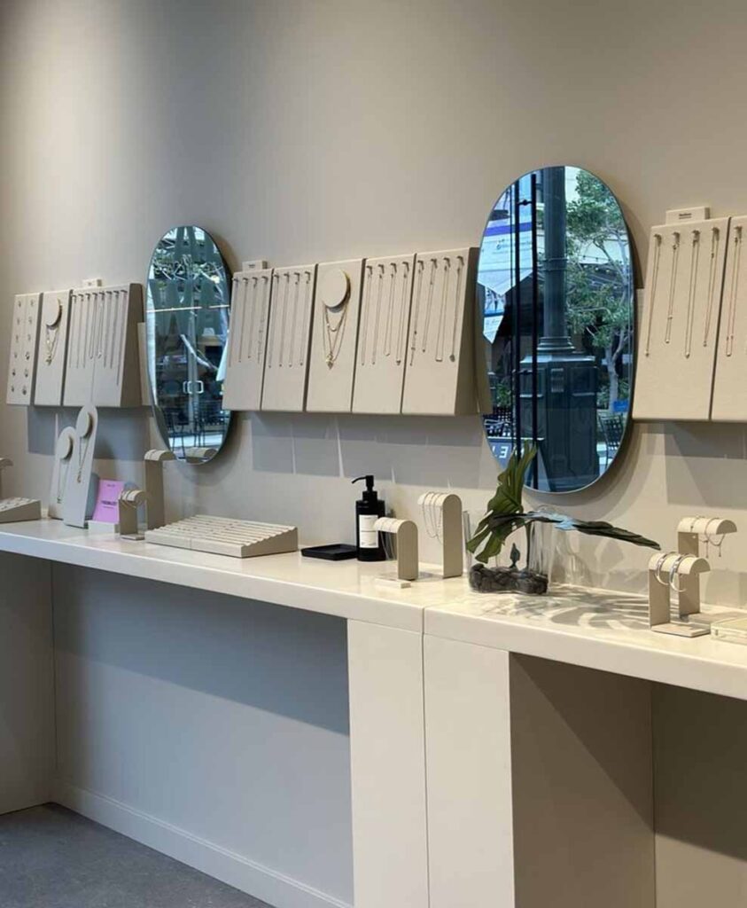 MOMA Construction Group in Glendale showcasing a custom built-in product display counter with upright minimalist necklace busts and sleek oval wall mirrors reflecting an outdoor pedestrian plaza.