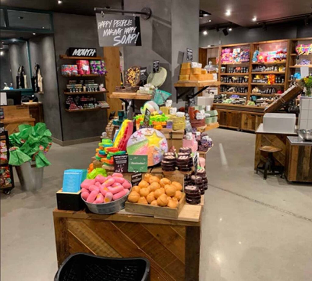 LUSH cosmetics shop interior featuring stacked wooden display tables with colorful bath bombs, soap bars, and a prominent chalk sign.