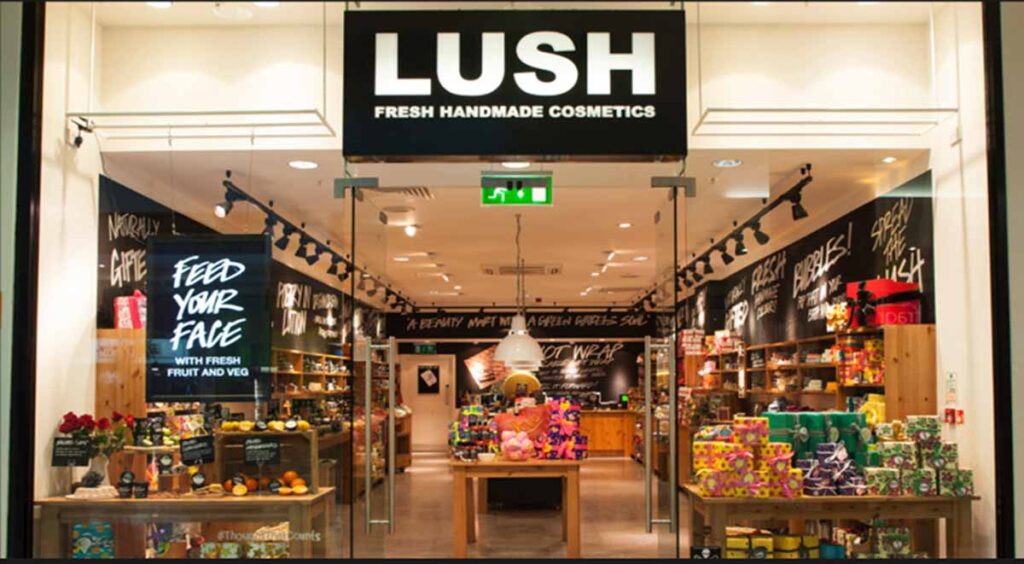 LUSH cosmetics storefront featuring a modern glass entryway, prominent illuminated branding signage, and vibrantly arranged product displays on rustic wood tables.
