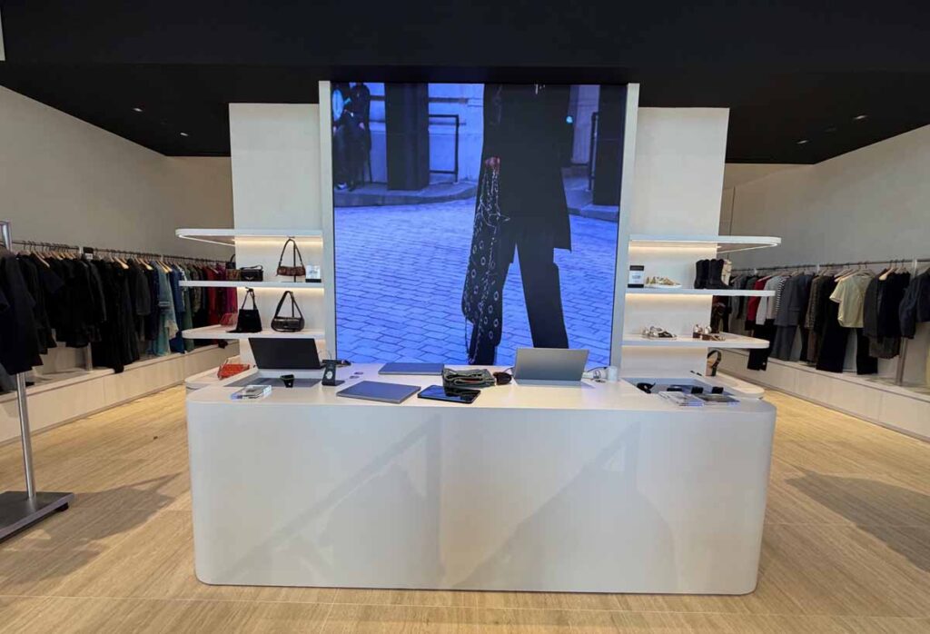 Sleek premium retail showroom featuring a large white geometric checkout counter, integrated baklit glass shelving, and a prominent freestanding digital display screen showing a fashion video.