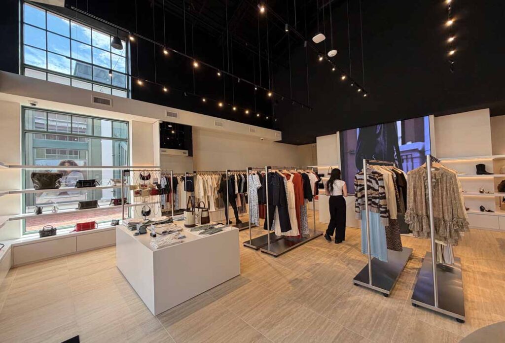 Spacious luxury fashion showroom featuring clothing racks, central geometric display islands, a large grid window overlooking an urban street, and overhead track lighting.