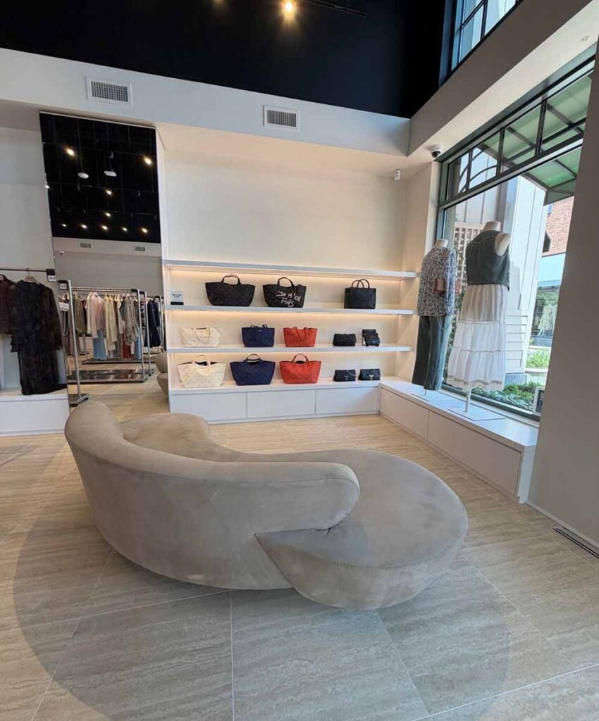 Upscale fashion boutique interior featuring an elegant curved plush sofa, built-in backlit wall shelving wiht luxury handbags, and a large display window overlooking an outdoor shopping district.