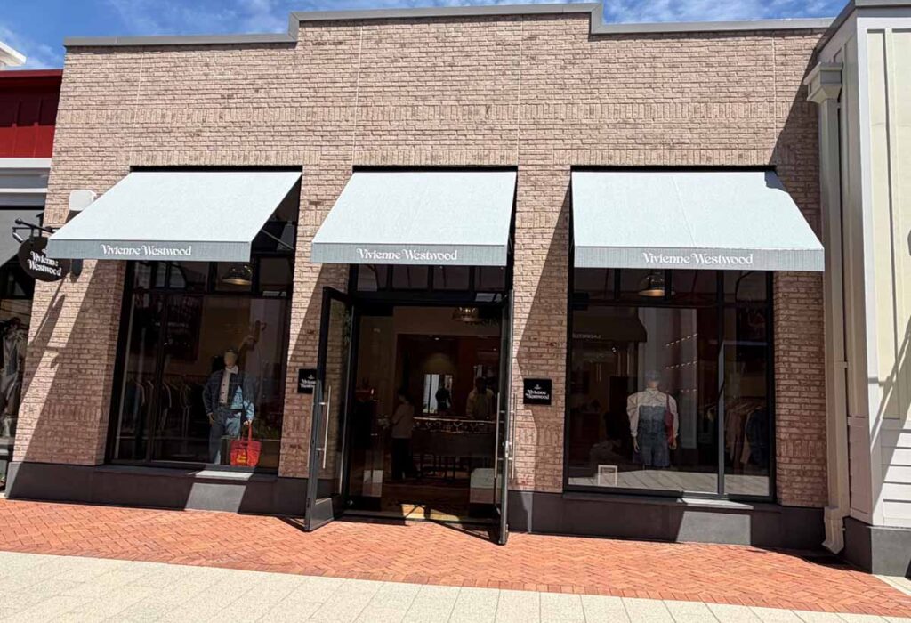 Upscale commercial retail storefront featuring a classic brick facade, large multi-pane glass display windows with prominent light gray Vivienne Westwood brand awnings, and a brick paver walkway.