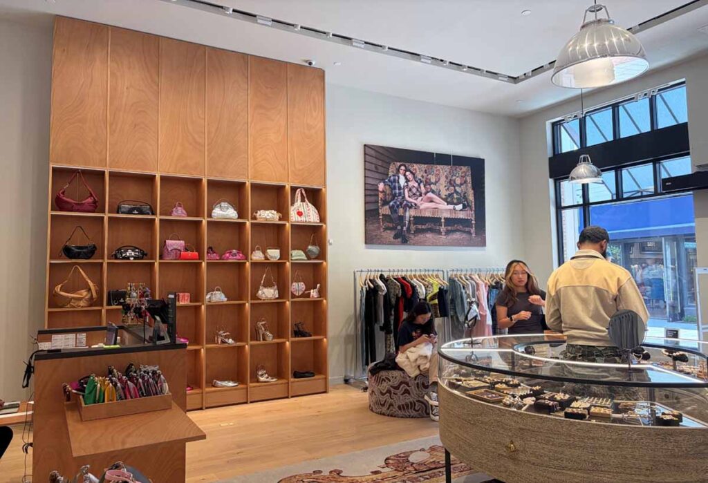 Upscale retail showroom featuring a massive floor-to-ceiling wooden shelving display filled with luxury handbags, alongside a curved glass jewelry presentation counter and ambient track lighting.