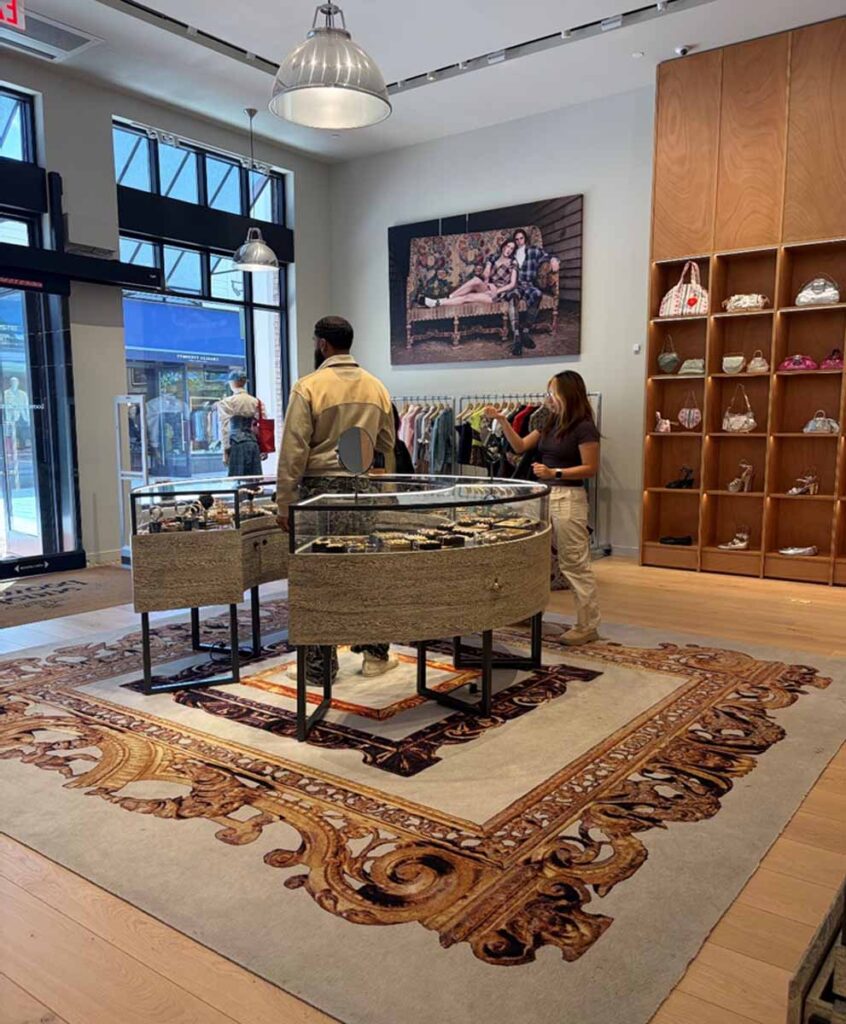 Upscale commercial clothing store interior featuring a circular wood-paneled jewelry display case, luxury apparel racks, integrated track lighting, and a large custom plush accent rug.