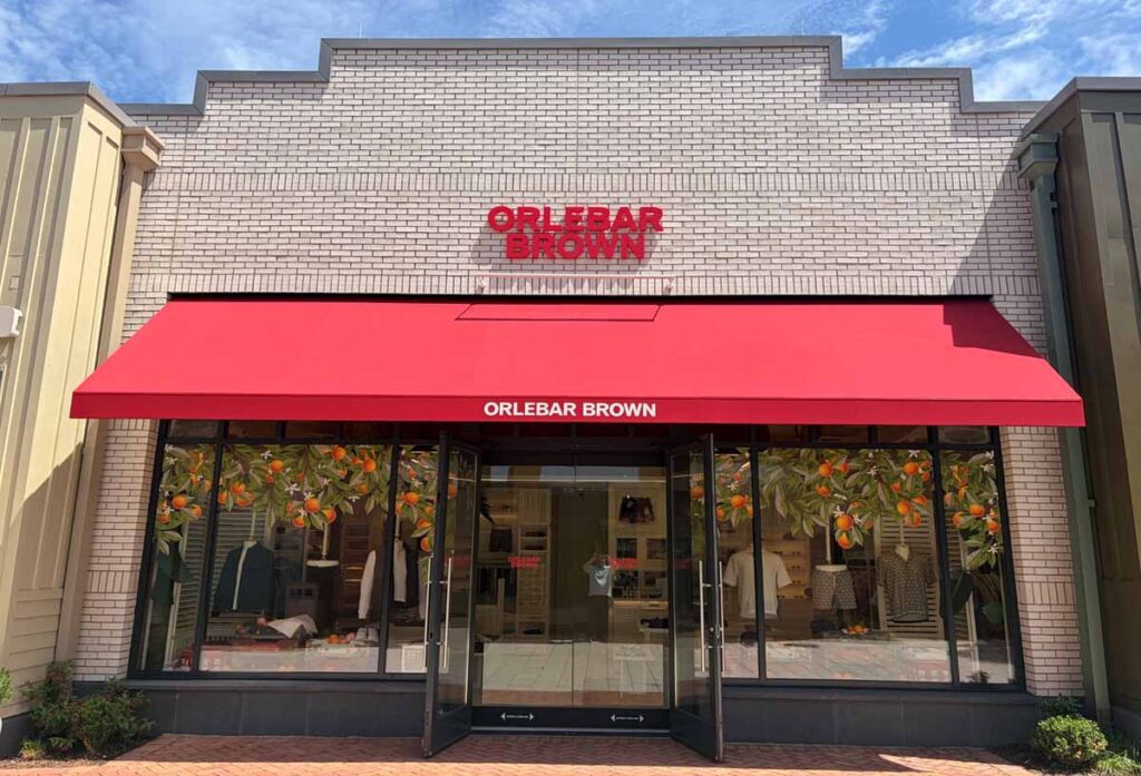 Upscale commercial retail storefront featuring a modern white brick facade, a vibrant red brand awning over glass entryway doors, and orange fruit tree window displays.