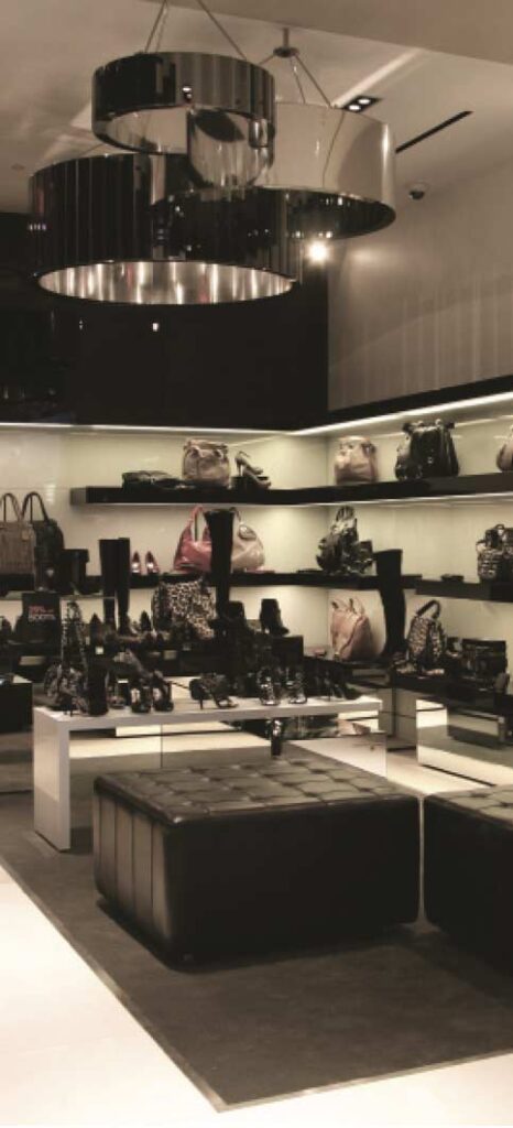 A high-end retail interior built by MOMA Construction Group for the Karen Millen luxury boutique, featuring custom-lit open shelving displaying designer handbags and shoes, modern circular tiered chandeliers, and a black tufted leather seating bench.