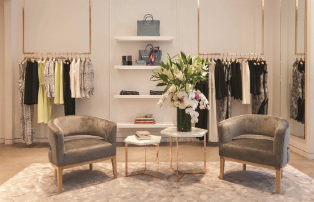 An elegant interior view of the Kimora Lee Simmons luxury retail boutique built by MOMA Construction Group, featuring a plush sitting lounge with two velvet armchairs, custom white shelving displays with gold accents, and clothing racks showcasing designer apparel.