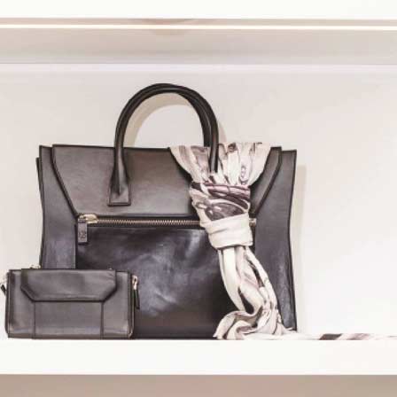 A close-up view of a luxury black leather designer handbag and matching clutch wallet displayed on a custom minimalist white floating shelf, built by MOMA Construction Group for the Kimora Lee Simmons boutique.