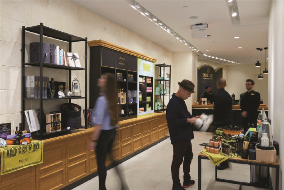 An interior showroom view of the Maille mustard boutique built by MOMA Construction Group, featuring a customer interacting at a sleek central tasting counter, extensive warm wood cabinetry stocked with premium jars, and industrial track lighting overhead.