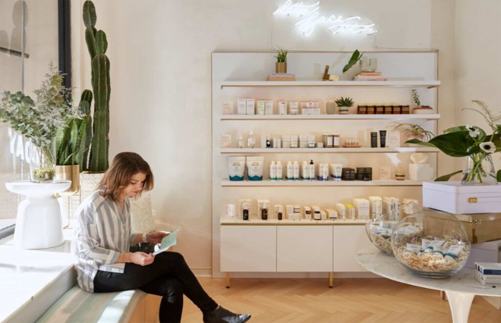 Modern boutique skincare showroom and salon interior featuring minimalist white built-in shelving, product displays, a herringbone wood floor, and a warm seating bench with a client.