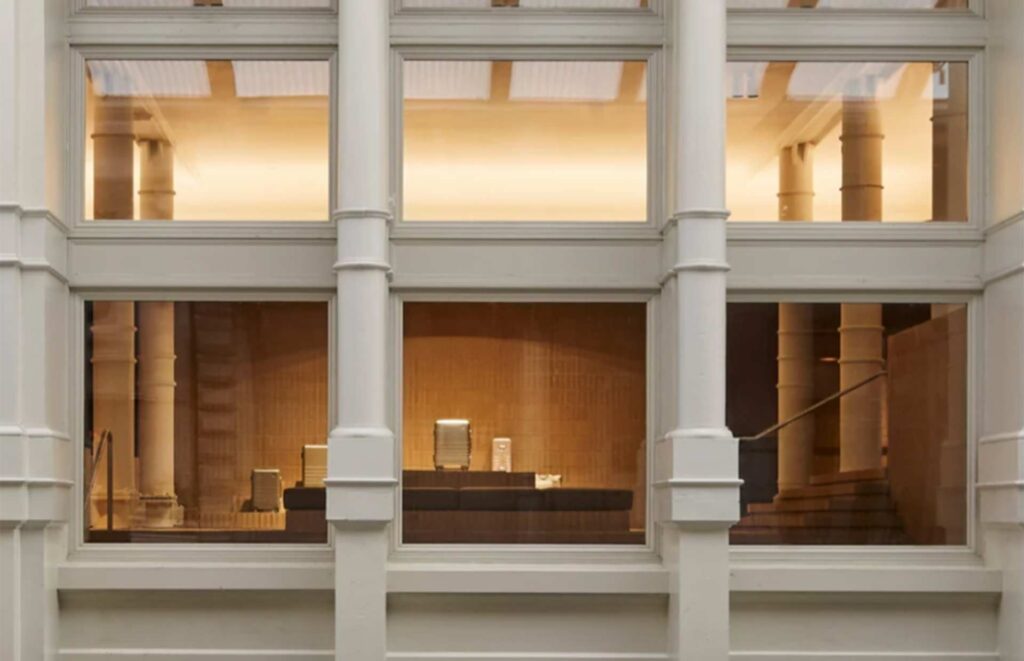 A street-level architectural view looking through the historic cast-iron window frames of the Monos retail showroom in Soho, built by MOMA Construction Group, showcasing a modern interior with prominent white structural columns and warm terracotta brick tile flooring.
