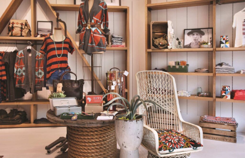 A detailed interior view of a custom apparel display area built by MOMA Construction Group for the Quicksilver retail boutique, featuring rustic wooden shelving stocked with folded clothing, apparel racks, a patterned wicker armchair, and styled coastal decor on a central wood display table.