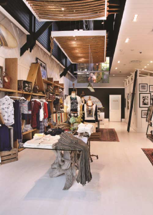 A wide interior view of the Quicksilver retail apparel boutique built by MOMA Construction Group, featuring custom wooden wall shelving filled with clothing, industrial ceiling panels wrapped in heavy decorative ropes, styled fashion mannequins, and glass-top display tables on a polished light gray floor.