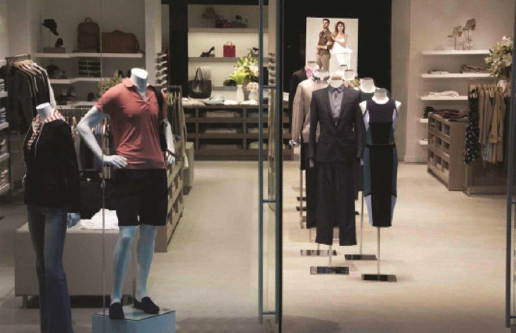 A high-end retail interior showroom built by MOMA Construction Group for Scoop NYC, featuring styled fashion mannequins behind structural glass partitions, custom built-in display shelving, and organized apparel racks under warm lighting.