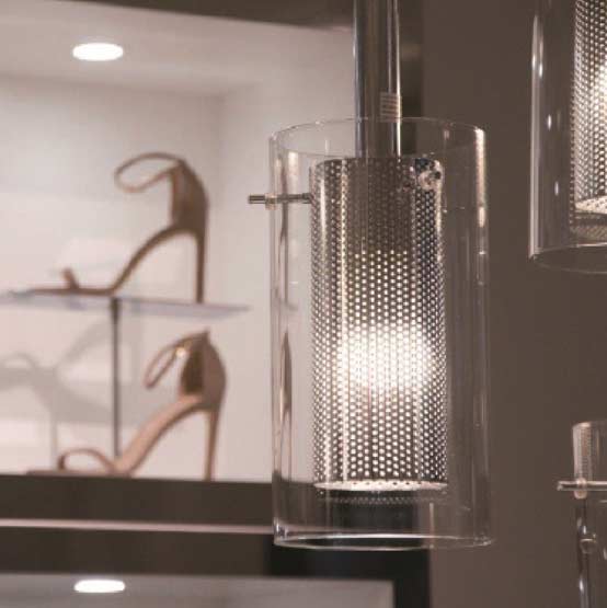 A close-up view of custom industrial cylindrical pendant lighting with perforated metal mesh inserts, installed by MOMA Construction Group in the Scoop NYC luxury retail showroom, with designer high heels displayed on glass shelving in the background.
