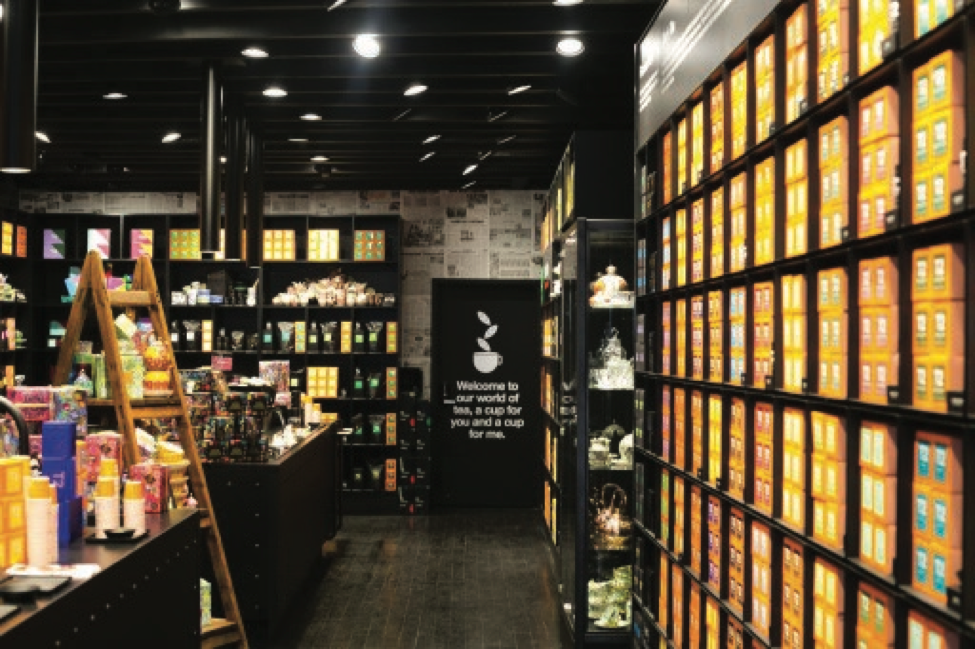 A dynamic interior view of the T2 Tea retail showroom built by MOMA Construction Group, highlighting extensive floor-to-ceiling black modular shelving filled with signature orange tea boxes, an open walkthrough layout, and a central display counter under industrial spotlighting.