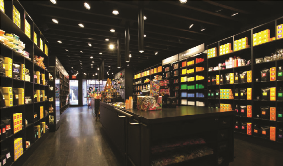 A wide interior view of the T2 Tea retail showroom built by MOMA Construction Group, featuring floor-to-ceiling matte black modular shelving units filled with brightly colored tea packaging, a dark wood central counter, and modern industrial track lighting.