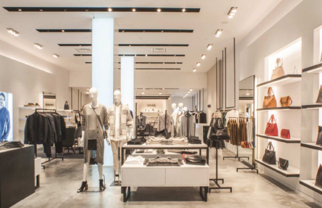 A wide interior view of the luxury Vince retail showroom in Soho built by MOMA Construction Group, featuring custom apparel display tables, minimalist structural columns, clothing racks with designer garments, and neatly arranged handbag shelving.